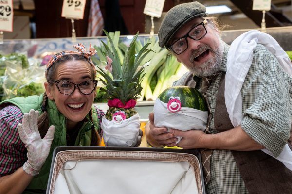 An image of a man and woman facing the camera and smiling. Between them there is a pineapple and a melon with dummies and a pair of eyes attached to them, and they are wrapped in white linen cloth.