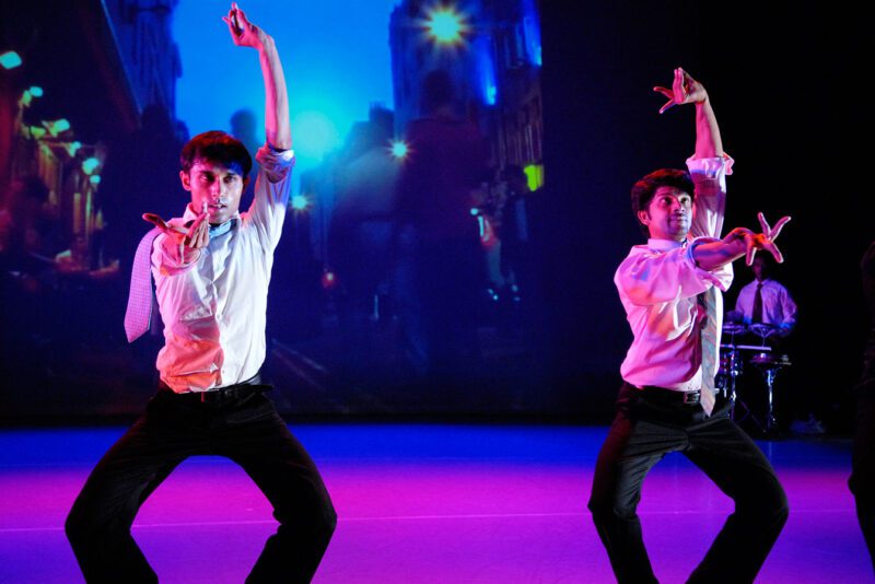 Two dancers are in an indian dance wearing business attire of black suit trousers, white cotton shirts with the sleeves rolled up and a tie. They are facing forward with their knees bent outwards. Their right arms are straight out in front of them with palms facing up. Their left arms are straight up above them with palms facing up. There is a city scene on the screen behind them and a drummer in the same outfit as the dancers.