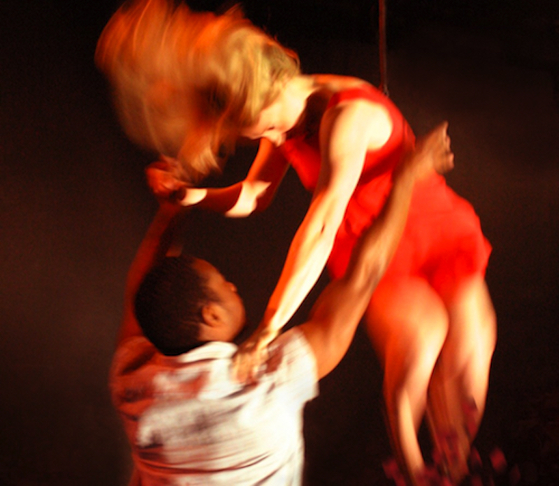 A dancer in a short sleeve white shirt holds onto their partner dancer who is wearing a red dress and flying through the air leaving a trail of red petals behind them.