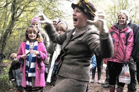 An image of a woman dressed in a brown jacket and hat makes a claw jesture with both hands. There is a small audience behind her.
