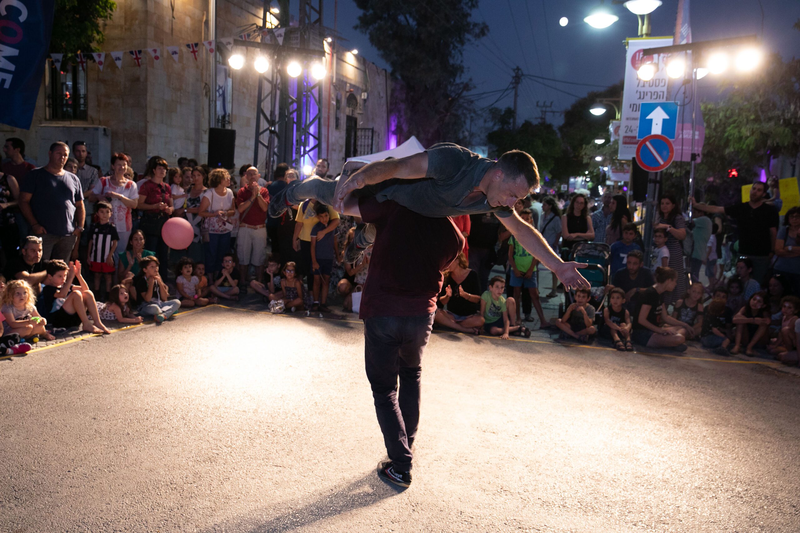 On the street at night time surrounded by a crowded audience, one performer is carrying another performer holding onto their hips on their left shoulder.