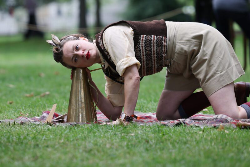 A performer is on their hands and knees listening to the ground through a brass eartrumpet they have placed on a blanket on the grass.