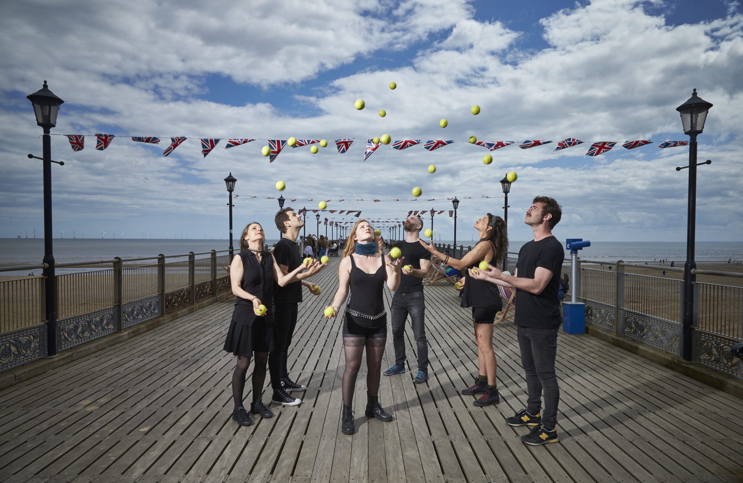 six people are juggling balls high into the air on a sea pier with bunting moving in the wind. The sky is mottled with clouds.