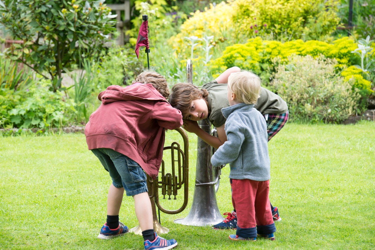 Three children are stood in a green space, two of them are bent over with their ears pressed to trumpets that are facing into the ground.
