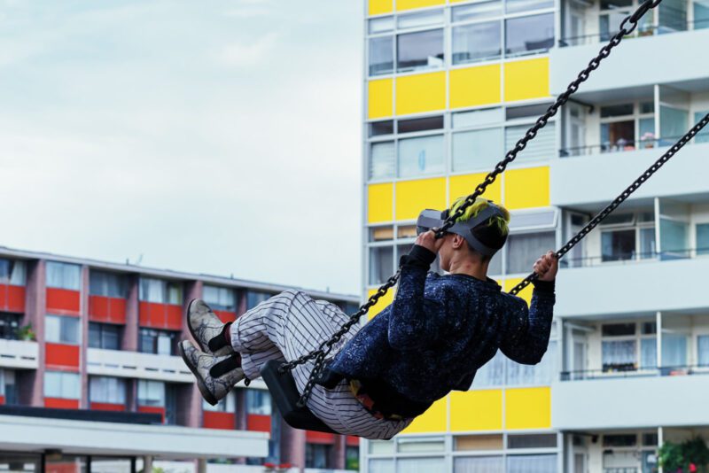 A person wearing a VR headset is mid air on a large swing amongst urban apartment buildings.