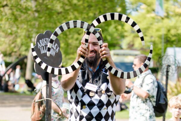 An image of a male performer facing the camera holding up two boomerang shaped objects with a black and white pattern.
