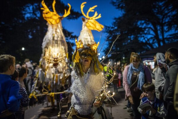 A night time image of a woman wearing a mask pulling along two stag shaped lanterns lit up from the inside.