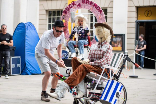An image of an audience members' interacting with mannequin which is sat on a garden chair. The mannequin is wearing a blonde wig, brown hat and brown trousers. The man is wearing a white T-shirt, beige shorts and sunglasses. There is a sign in the background saying 'Buck a Brenda'.
