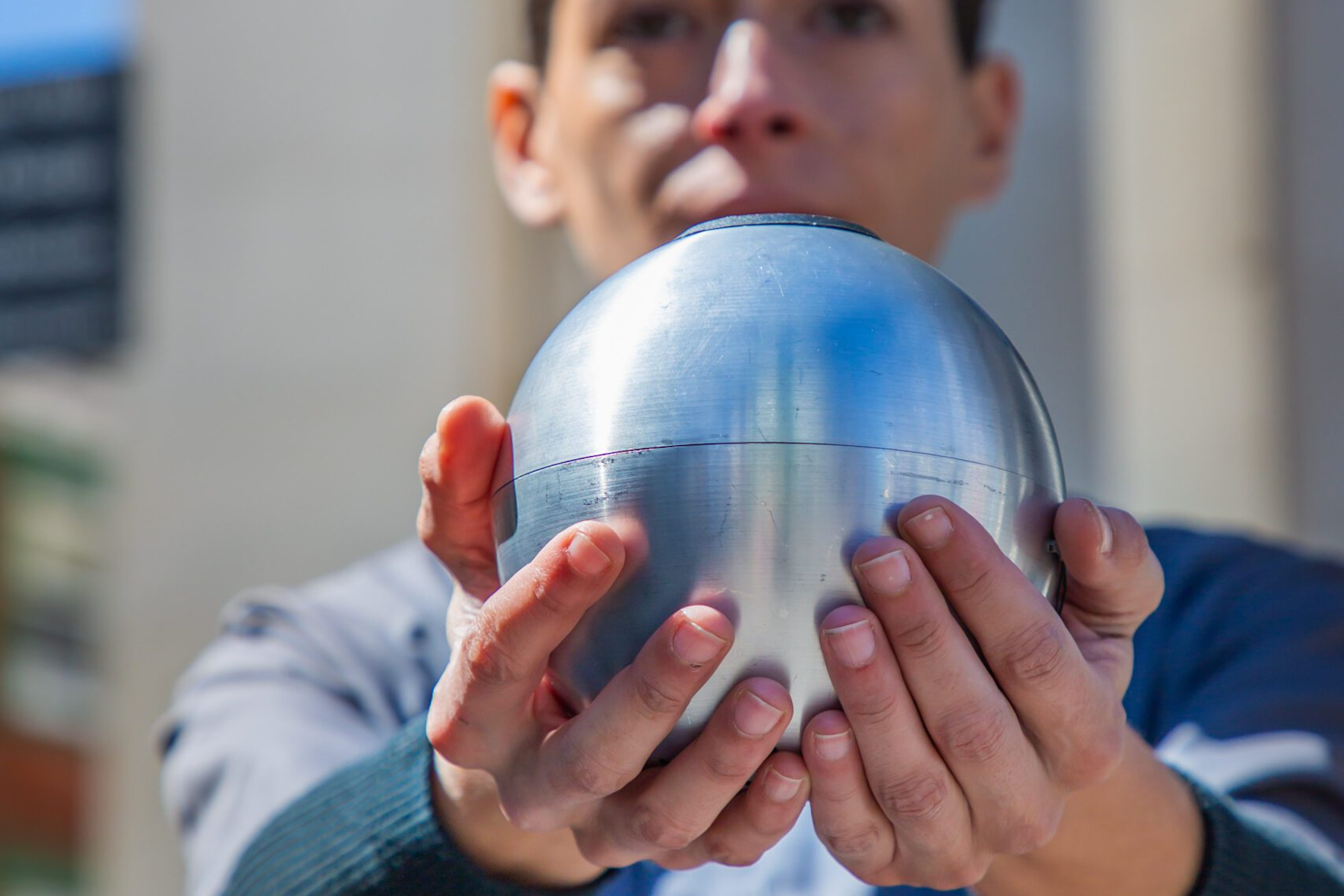 A person standing outside in the sun is holding a metal ball cupped in their hands that are out in front of them at shoulder height.