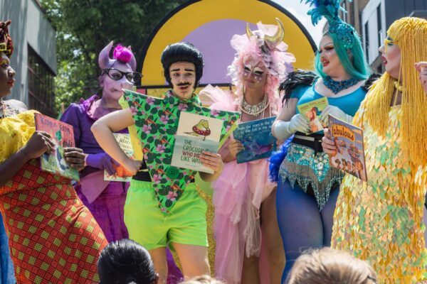 An image of six drag queens each carrying a children's book.