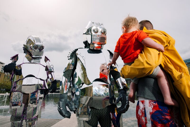 An image of two people dressed in metallic costume greeting a man carrying a small child.