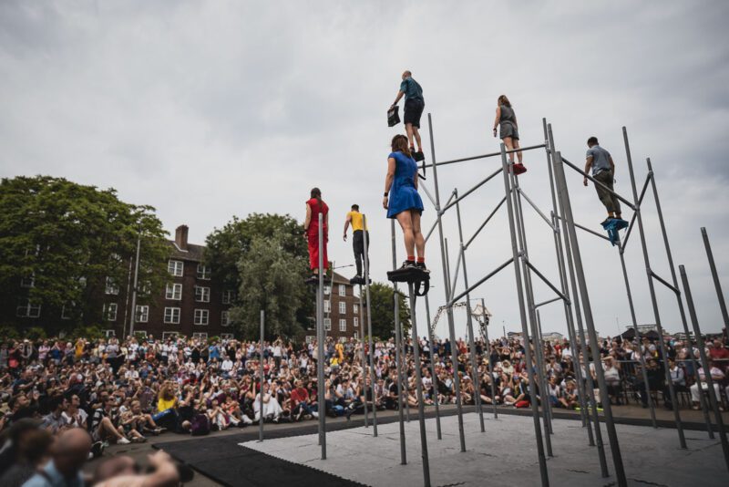 An image of a group of performers stood at various points on top of a large steel frame structure. There is a crowd of people below them.