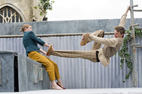 An image of a male and female performer. He is hanging from scaffolding and she is pulling his leg.