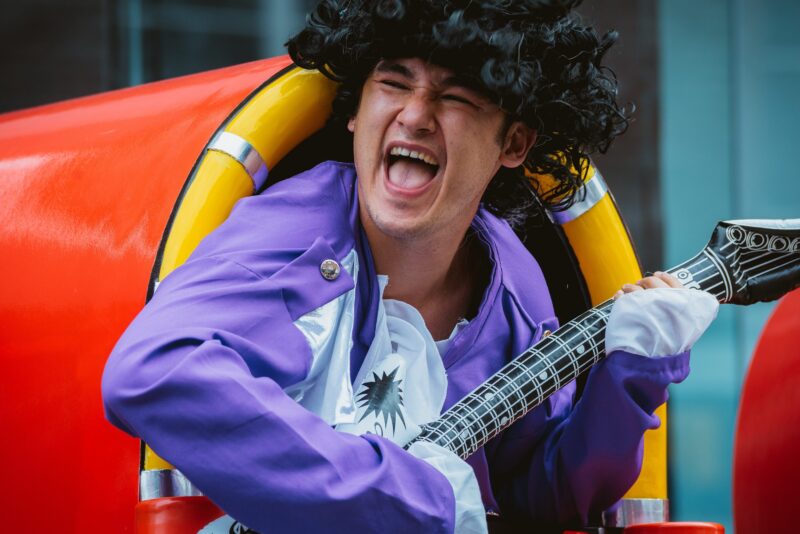 An image of a man peering out of a jukebox wearing a black wig, a purple shirt and playing an inflatable guitar.
