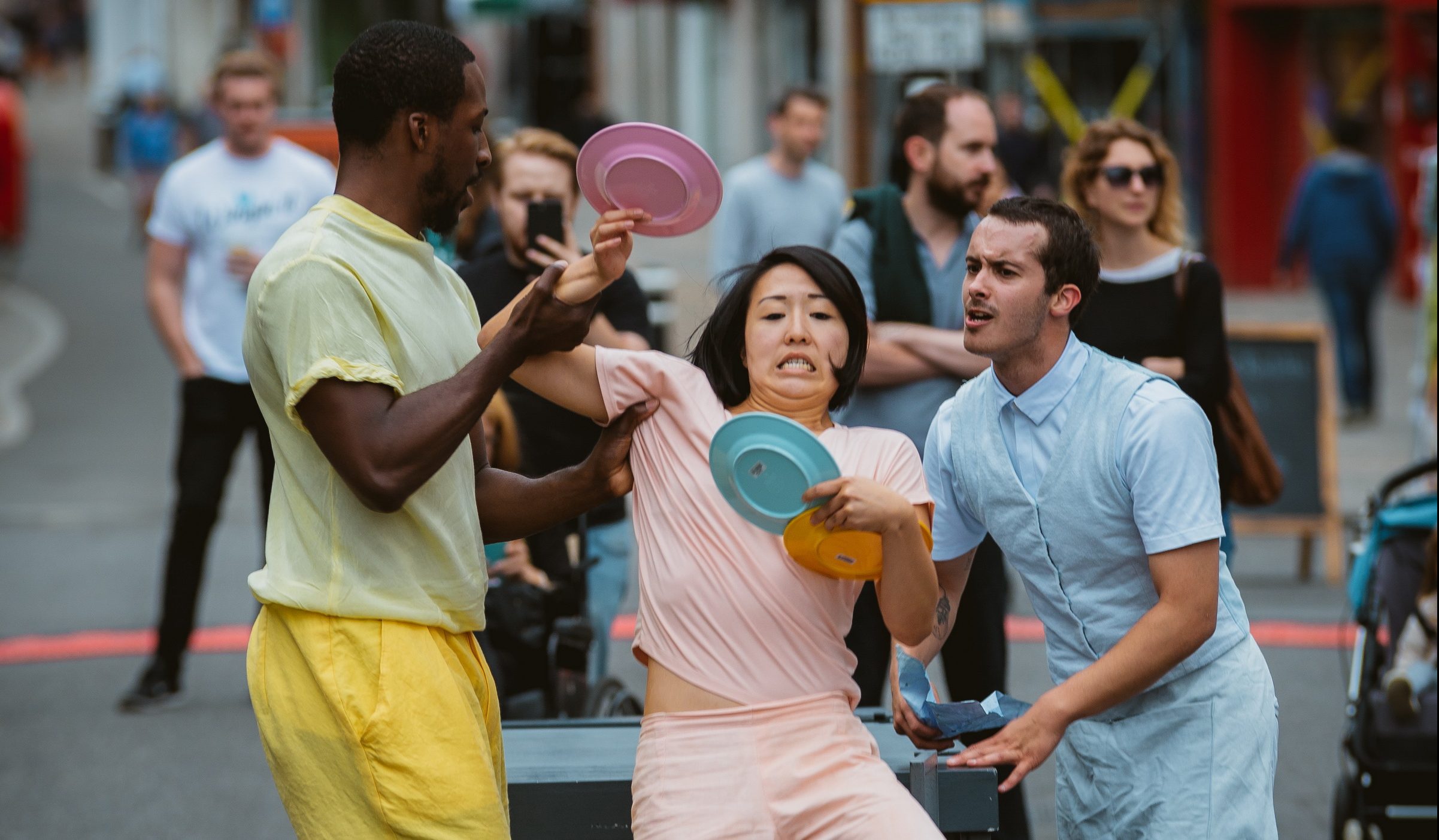 An image of two male performers and one female. The two males are holding up the female who is losing her balance whilst holding three plastic plates. They are all wearing pastel coloured clothing.