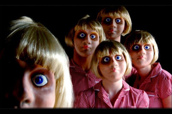 An image of five doll like faces with wide blue eyes and blonde wigs with a dark background.