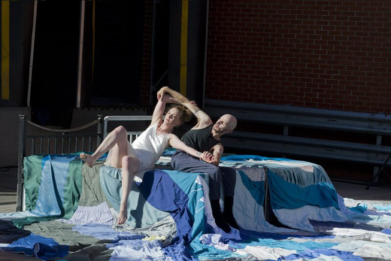 An image of a male and female performer clasping hands and sat on a large makeshift bed with a patchwork quilt draped over it.