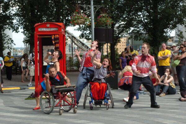 An image of a group of dancers, some are wheelchair users, performing and making gestures in front of a red phone box.