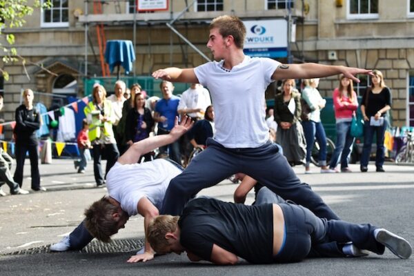 An image of three male performers. Two of them are on the floor and one of them is stood between them in a warrior like stance. They are wearing grey jogging bottoms and t-shirts.