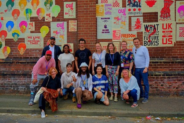 An image of a group of people stood and sat in front of a brick wall with various posters displayed.