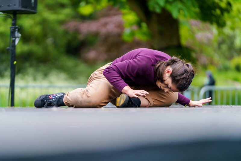 An image of a female dancer crouched down on the floor with her head bowed. She is wearing a purple top and beige trousers.