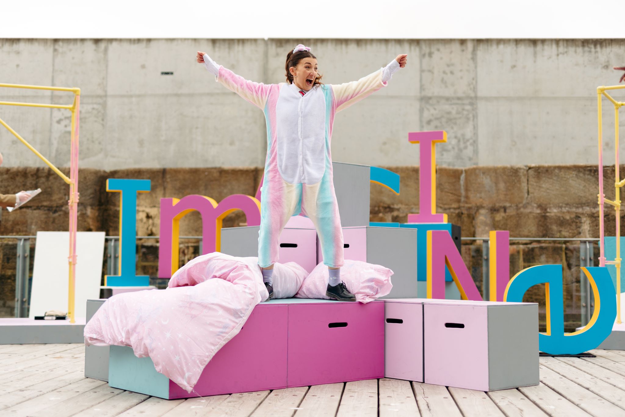 Atop pastel-coloured boxes draped in a pink duvet, a performer in a pastel-coloured onesie sings with her arms raised up.