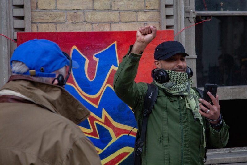 An image of a man with his fist in the air. He is wearing a scarf wrapped around his face and a green jacket, and holding a phone in his other hand. There is a man in front of him with his back to the camera wearing a blue baseball cap.
