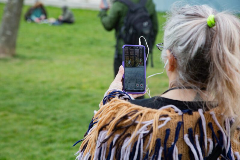 An image of a woman with her back to the camera pointing her phone at something with a grassy background.