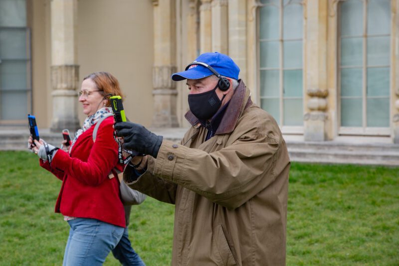 An image of a man and woman taking a photo or filming something with their mobile phones. The man is wearing a blue baseball cap, face mask and brown jacket, and the woman is wearing a red jacket.