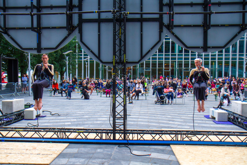 An image of a stage with a male and female trapeze artist hanging from a fixed point with a crowd of people watching in the background.