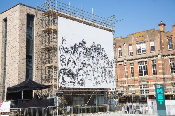 An image of a billboard fixed against a building with scaffolding behind it. The billboard has a white background and depicts a pencil drawing of human faces.