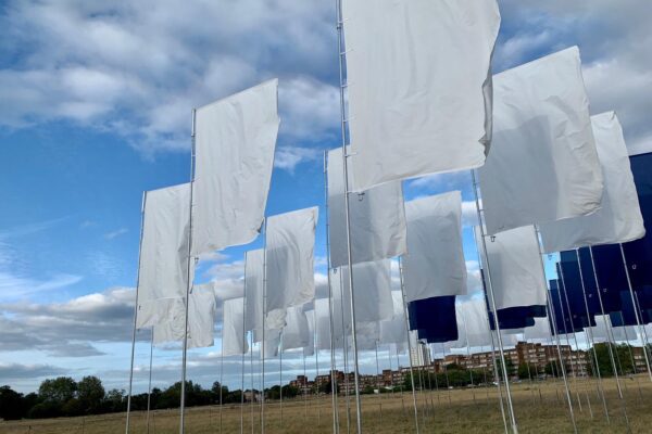 An image of rows of white flags fixed into the ground in a field with a blue sky cloudy background.