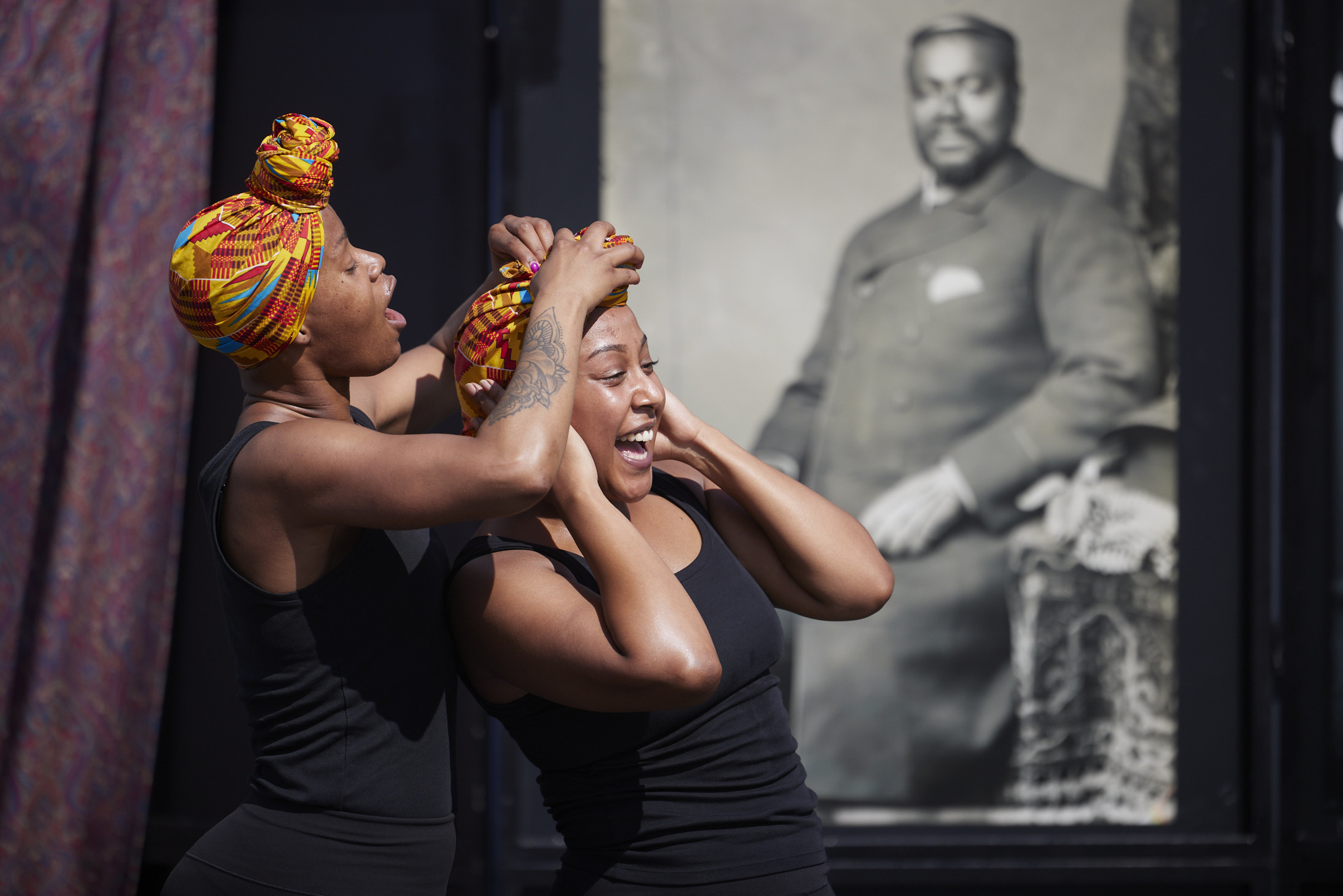Two performers are interacting on stage. One is wearing a colourful head wrap while fixing the other's head wrap for them.