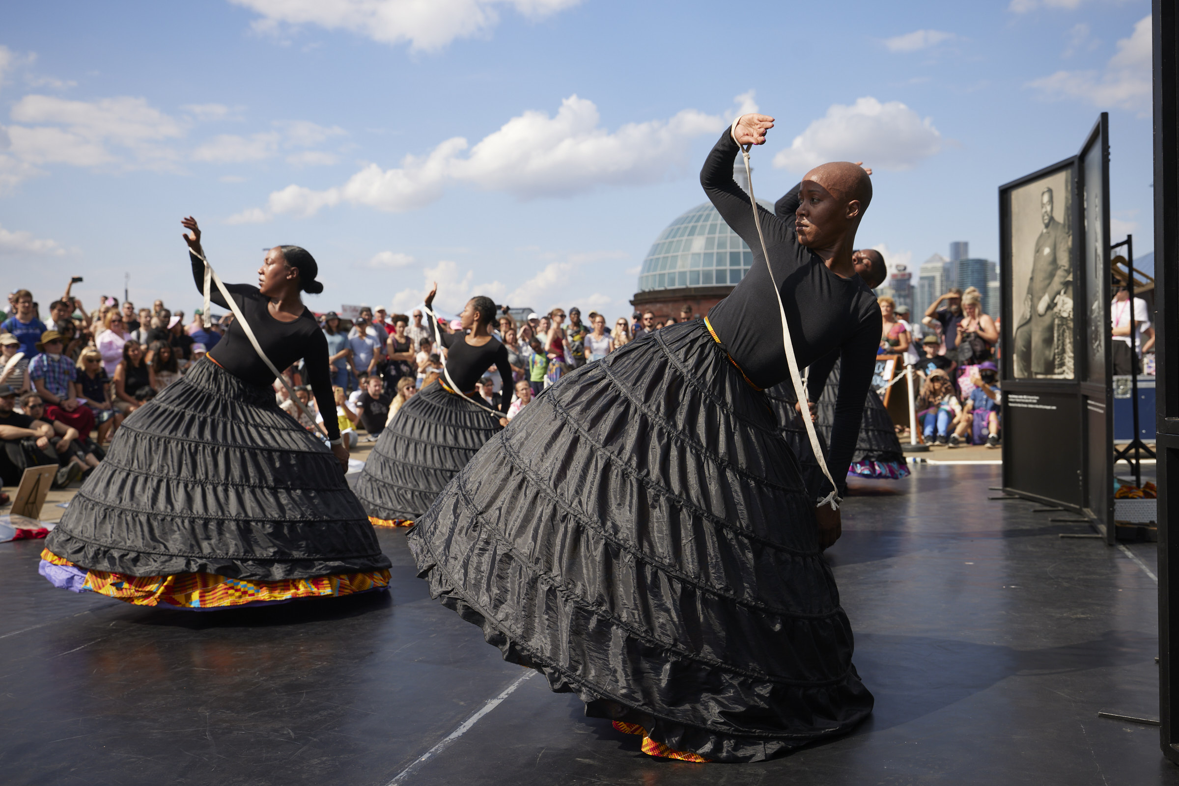 Performers are wearing black tops with large black cage skirts that are on top of colourful cage skirts underneath. All of them are bound by a rope from wrist to wrist. It is a sunny day and they are surrounded by a crowded audience.