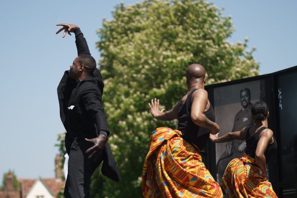 We see one male and two female black dancers with their backs to the camera. They are wearing black and yellow clothing and there is a tall tree and blue sky background.