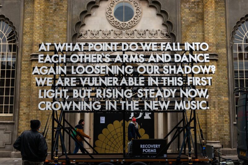An image of a large body of text fixed against the doorway of an ornate building. The text reads 'At what point do we fall into each other's arms and dance again loosening our shadow? We are vulnerable in this first light, but rising steady now, crowning in the new music'.