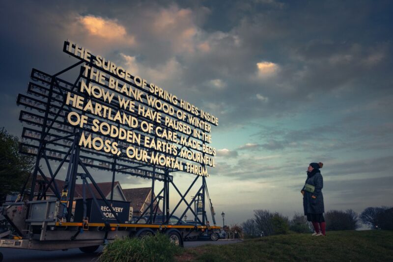 A night time image of a large body of text fixed onto a framed structure. There is a person reading it to the right. The text reads 'The surge of spring hides inside the blank wood now we have paused in the heartland of care, made beds of sodden earth's mournful moss, our mortal thrum.'