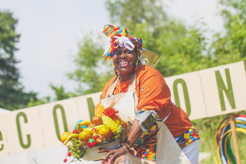An image of a black woman wearing traditional African dress and headdress. She is wearing an apron and holding a fruit bowl.