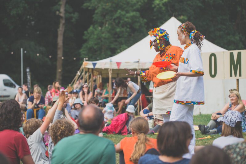 We see two female performers stood up in front of a crowd who are sat down and reaching out to them.