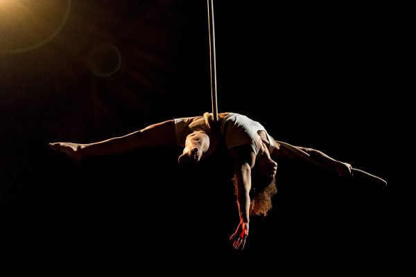 An image of a trapeze artists suspended from the ceiling wearing dark clothing against a dakr background.