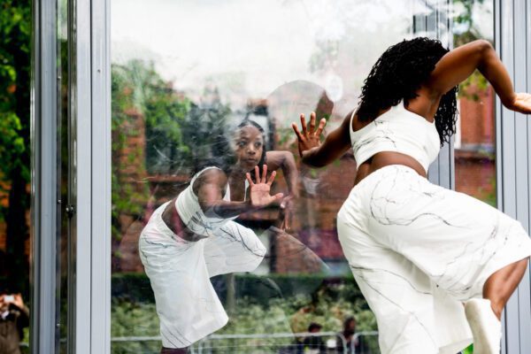 An image of two female black dancers facing one another with their hand on a glass wall. They are both all dressed in white.