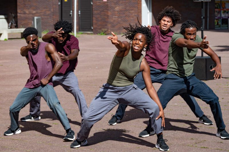 An image of four black male dancers and one female. They are all wearing blue pants and purple and green t-shirts.