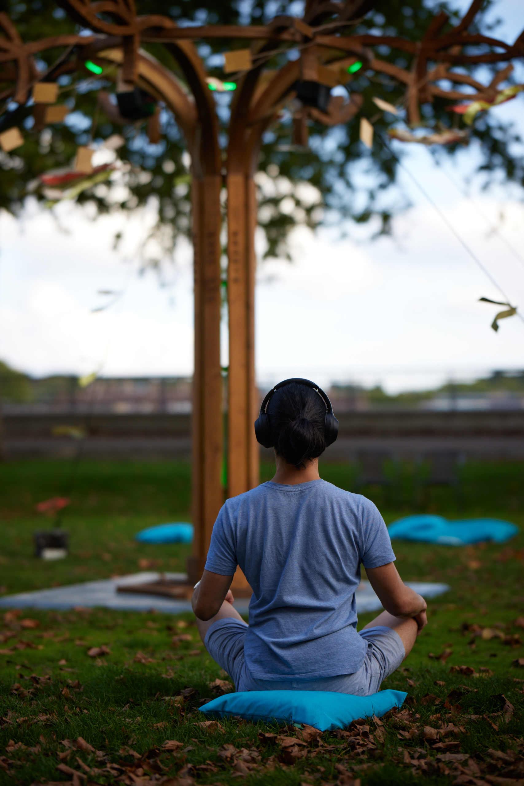A person wearing headphones listening to a sound installation sits in a meditative position on a blue cushion on the floor. They are looking into the distance and sat beneath a tree whilst the sun sets. They are listening to Final Farewell by Tara Theatre at GDIF 2022.