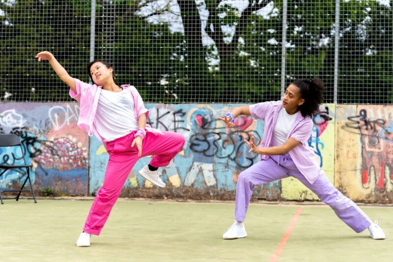 Two dancers are actively moving between dance poses. They are in front of a fence that is graffitied concrete at the bottom and wire at the top showing the trees behind.