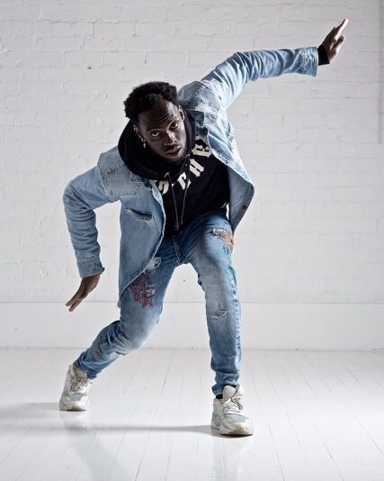 Chad Taylor poses, as if in mid-dance with his knees slightly bent and his arms bent in different directions. He is in a white room. He is wearing a denim jacket and ripped jeans, with a black Trapstar hoodie under his jacket.