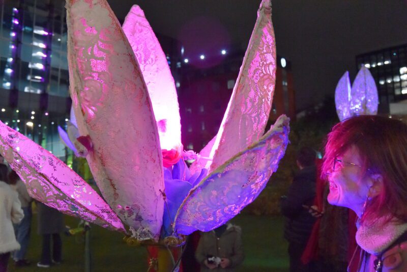 Outdoors at night, someone is smiling and looking up close at a big robotic flower, which has petals made out of lace and bent metal wire is lit by a pink light.Tucked inside is purple felt that resembles more petals.