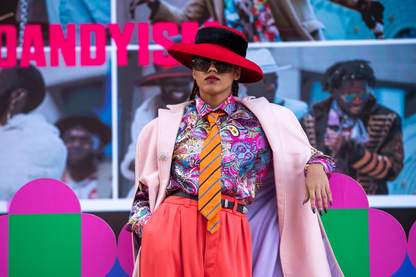 A performer poses wearing bold and colourful clothes: a red hat with black faux fur band; sunglasses; a multicoloured, mostly pink, paisley silk shirt; a pastel pink overcoat with lilac lining and a silver flower brooch pinned on the lapel; orange striped tie; and blood orange high-waisted trousers with a black belt.