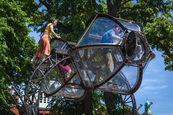 An image of a large egg timer like structure that moves on an axis. There is a performer inside the pod closest to the camera and a performer stood on the pod behind, with a blue sky and green trees in the background.