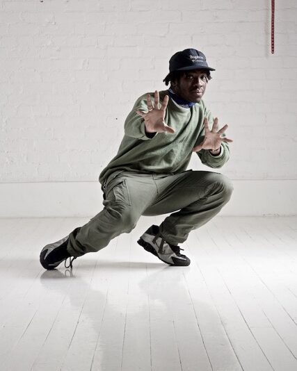 Ziggy Taylor poses, crouching with his weight on his left knee and his arms oustretched pointed at the camera. He is wearing a black cap with khaki sweatshirt and cargo trousers.