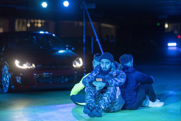 An image of three man sat in a circle with their backs to one another. The man facing the camera is wearing army style clothing and there is a car behind them with it's lights on.